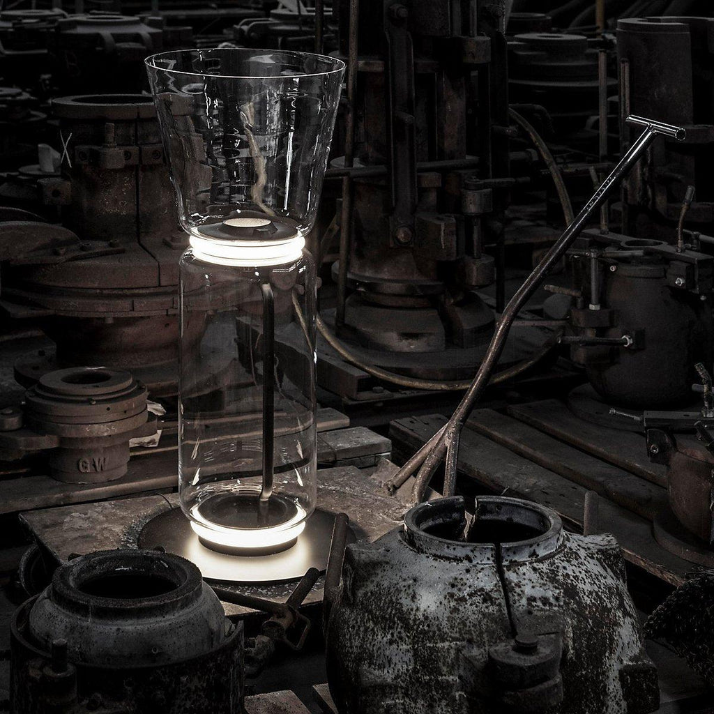 A Clérachic glass floor lamp with a cone shade standing among old iron machinery in a dark industrial space.