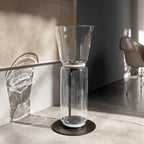 Clérachic glass floor lamp with a wide cone shade in a bare concrete room, with a chair and glass shadows on the wall.
