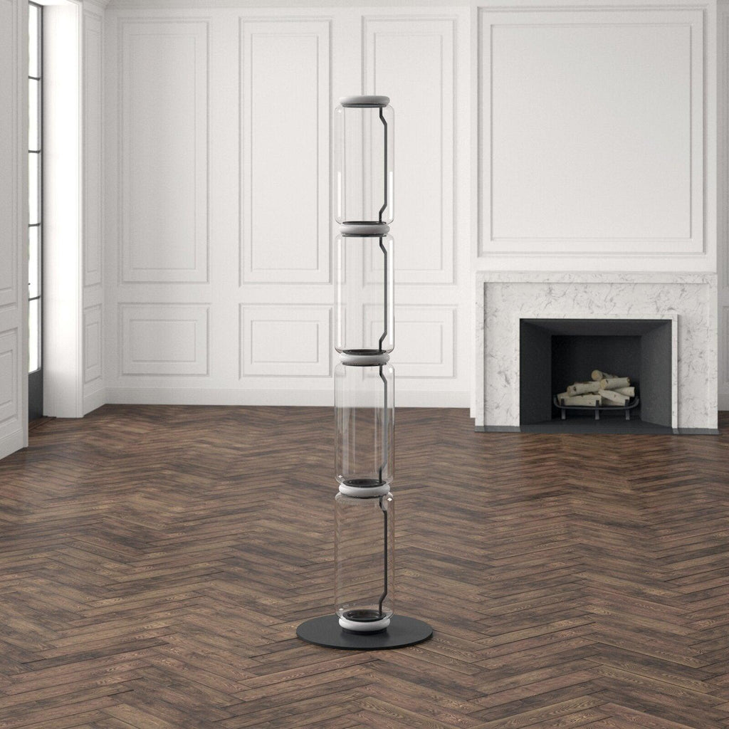 Tall unlit Clérachic glass floor lamp on a herringbone floor in a white panelled room, black connectors showing between the glass sections.