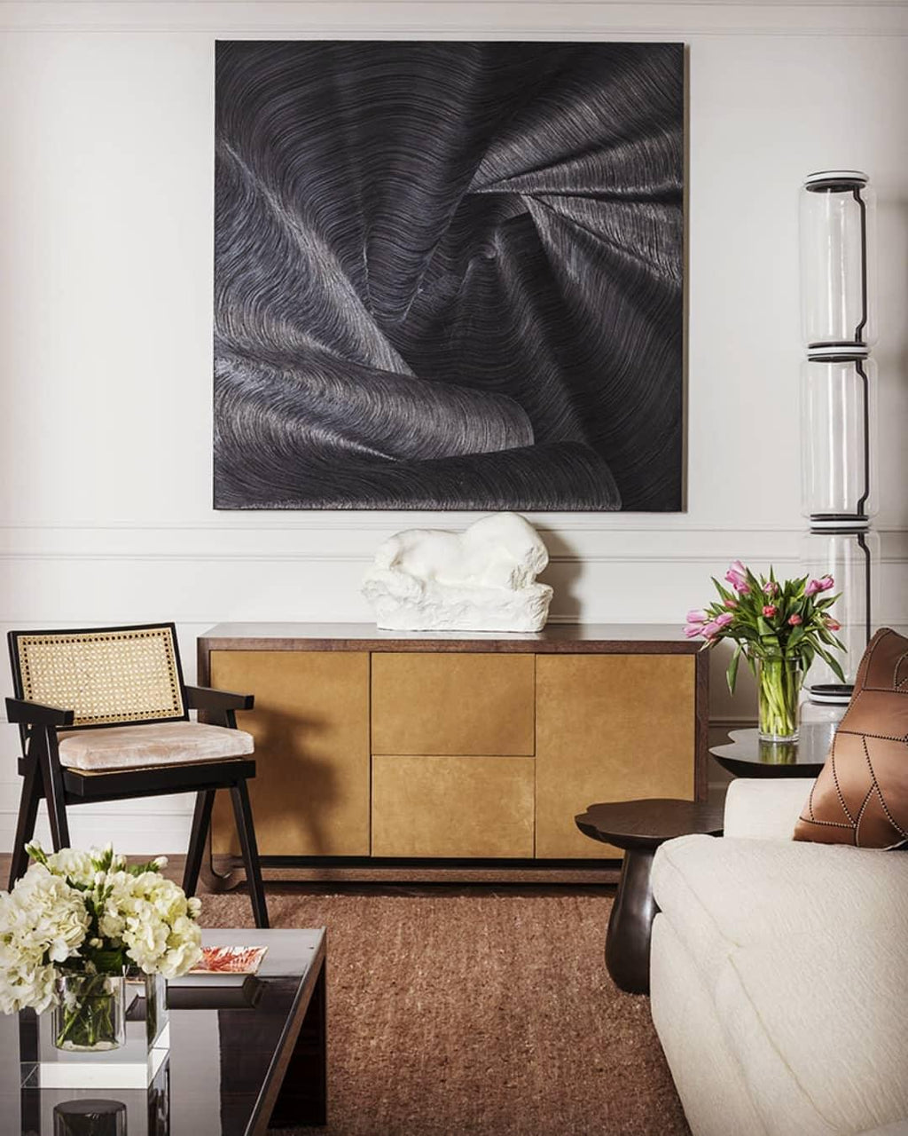 Clérachic glass floor lamp at the edge of a sitting room, beside a large dark artwork, a wooden sideboard and a bunch of tulips.