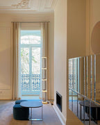 Slim Clérachic glass floor lamp near a tall window with long curtains, beside a navy stool and a low round table.