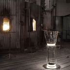 A Clérachic glass floor lamp with a cone shade in an old industrial building, with timber-clad walls and a lit window opening.
