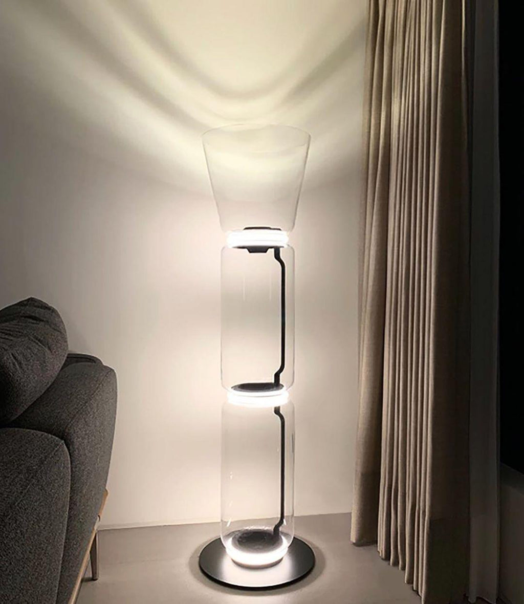 Clérachic glass floor lamp with a cone shade lit in a corner beside a grey armchair and a long taupe curtain.