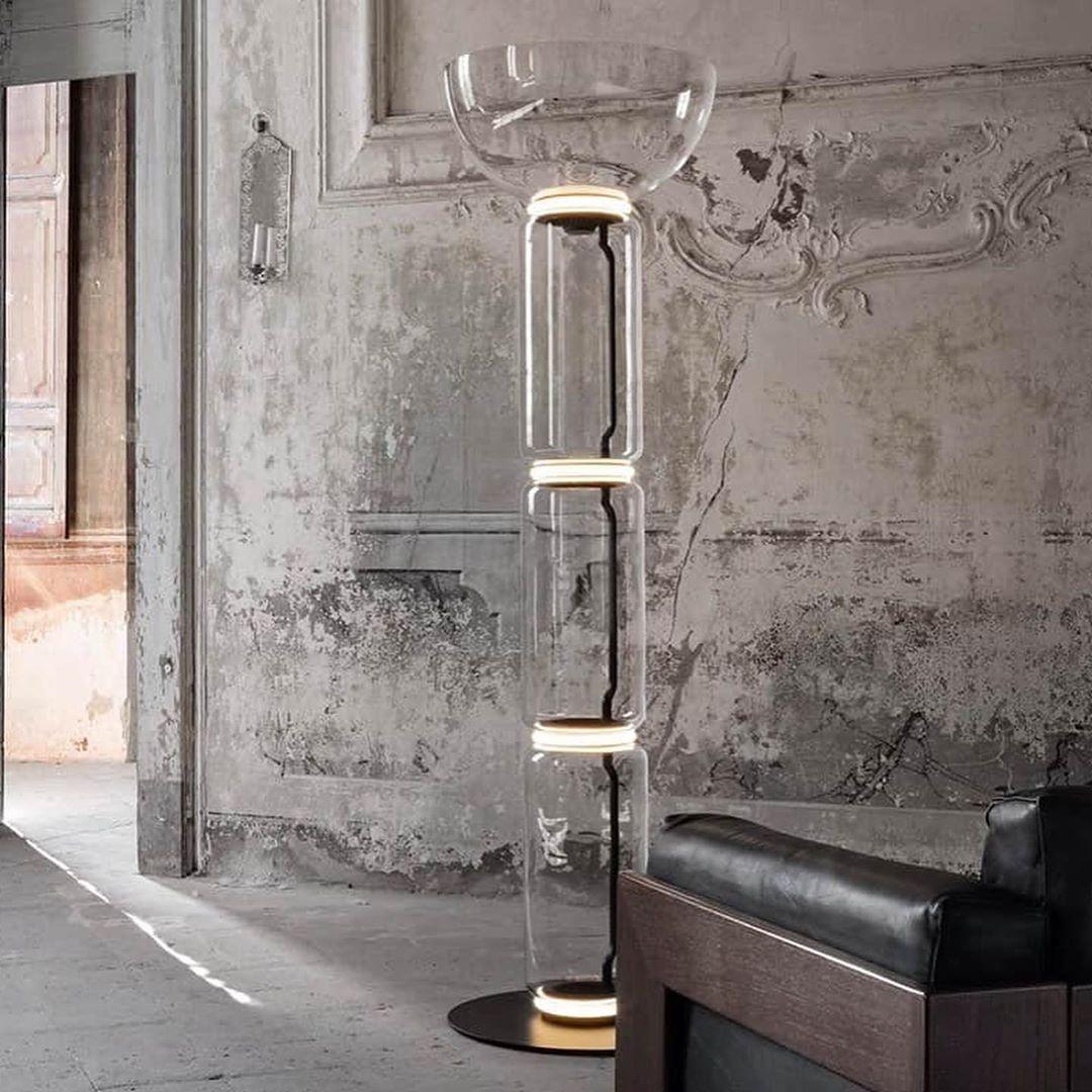 A Clérachic glass floor lamp with a bowl shade against a crumbling plaster wall, with a dark leather armchair at the right.
