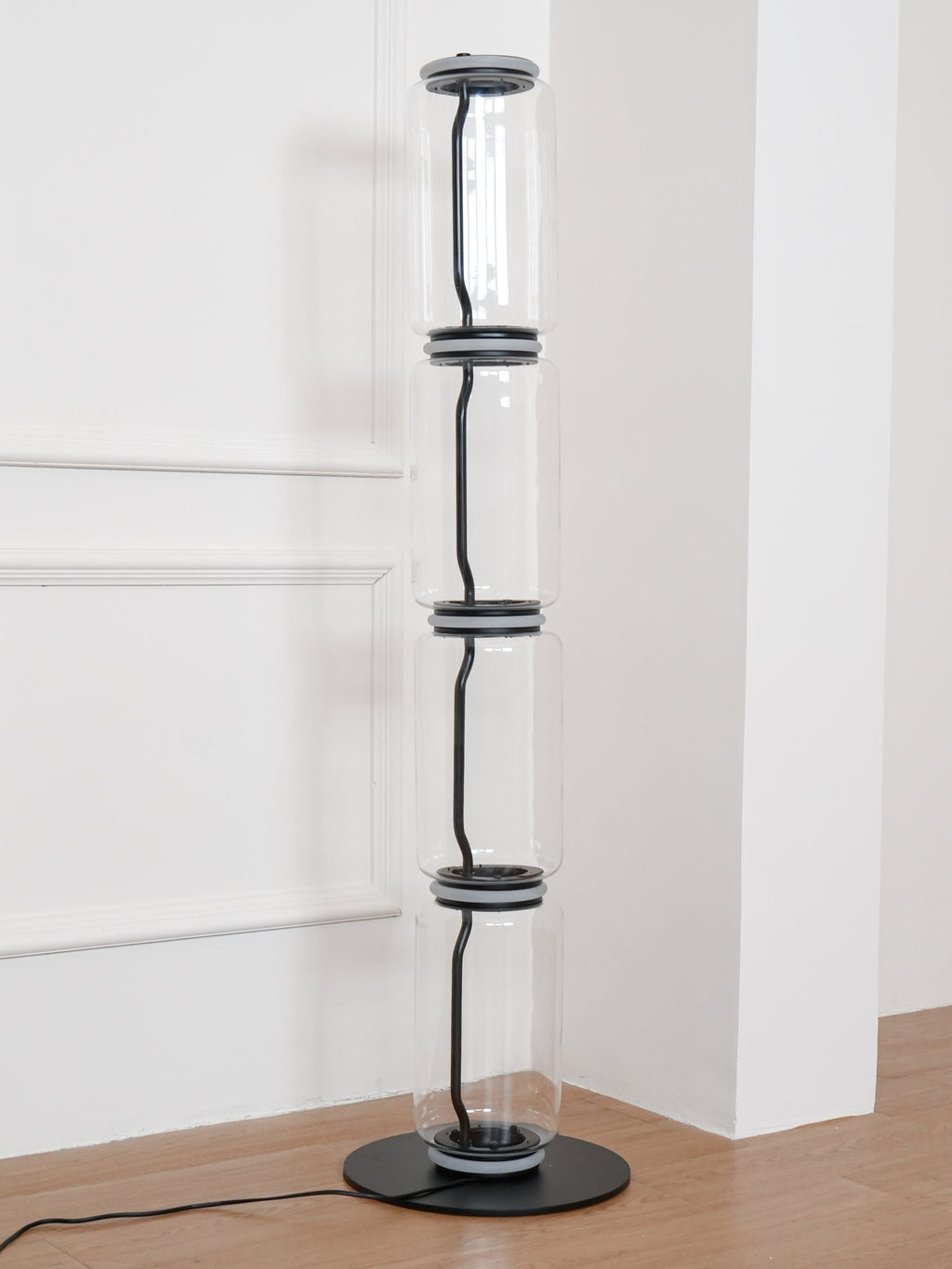 Unlit Clérachic glass floor lamp in a white corner, a black cord running through four clear cylinders above a round black base.