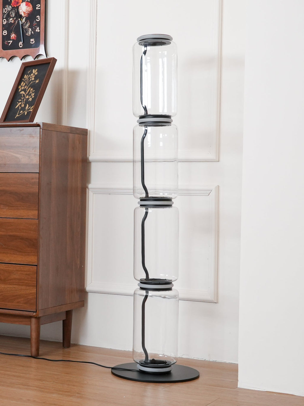 Unlit Clérachic glass floor lamp beside a walnut chest with a framed floral picture, against a white panelled wall.