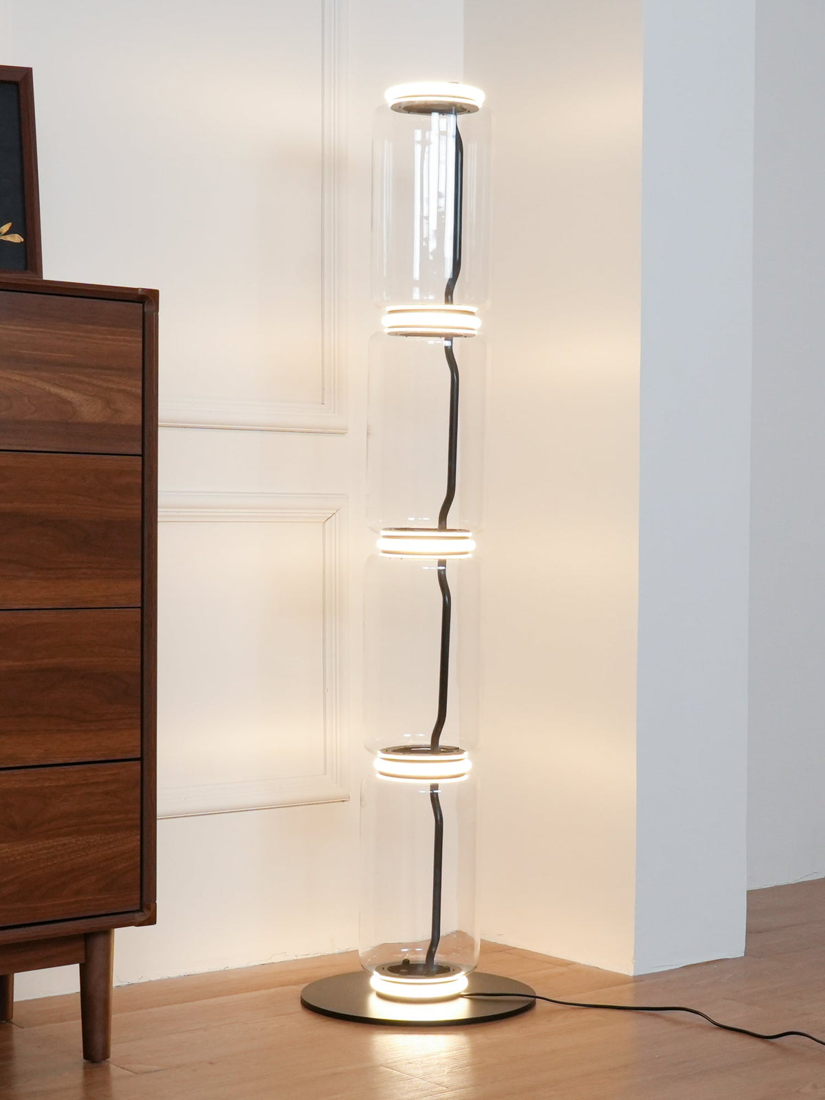 Clérachic glass floor lamp glowing warm in a corner with a walnut chest of drawers, light rings stacked up the glass.