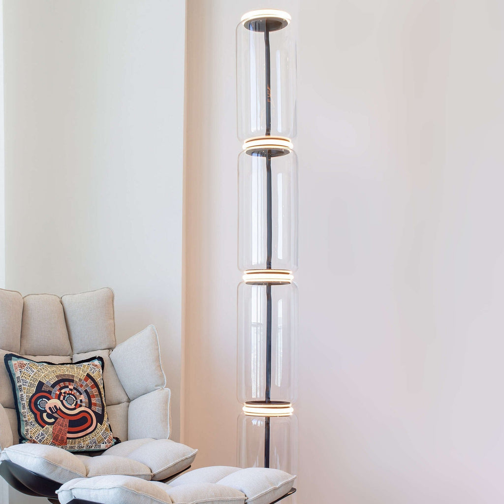 Clérachic glass floor lamp lit beside a cream lounge chair with a patterned cushion, in a pale pink-white room.