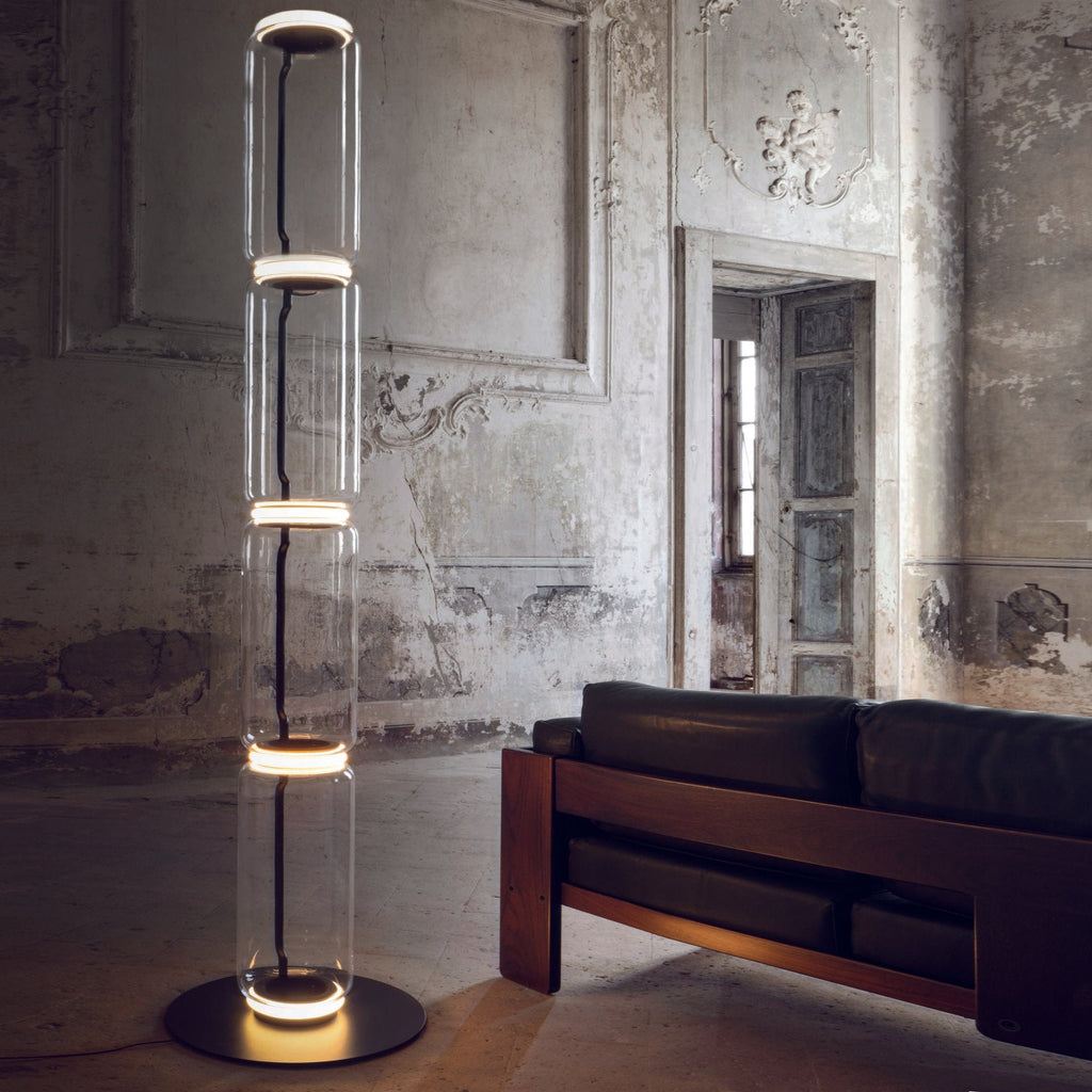 Clérachic glass floor lamp lit in a room with ornate plaster walls, beside a dark leather sofa on a stone floor.