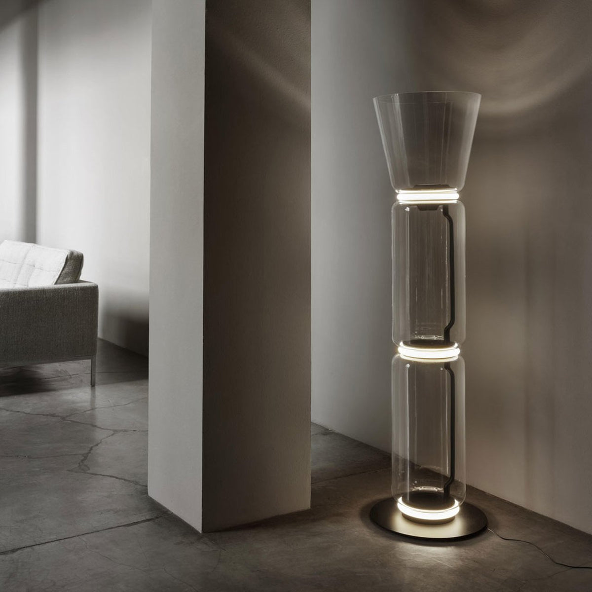 Clérachic glass floor lamp with a cone shade glowing on a concrete floor beside a plain pillar, with a sofa behind it.