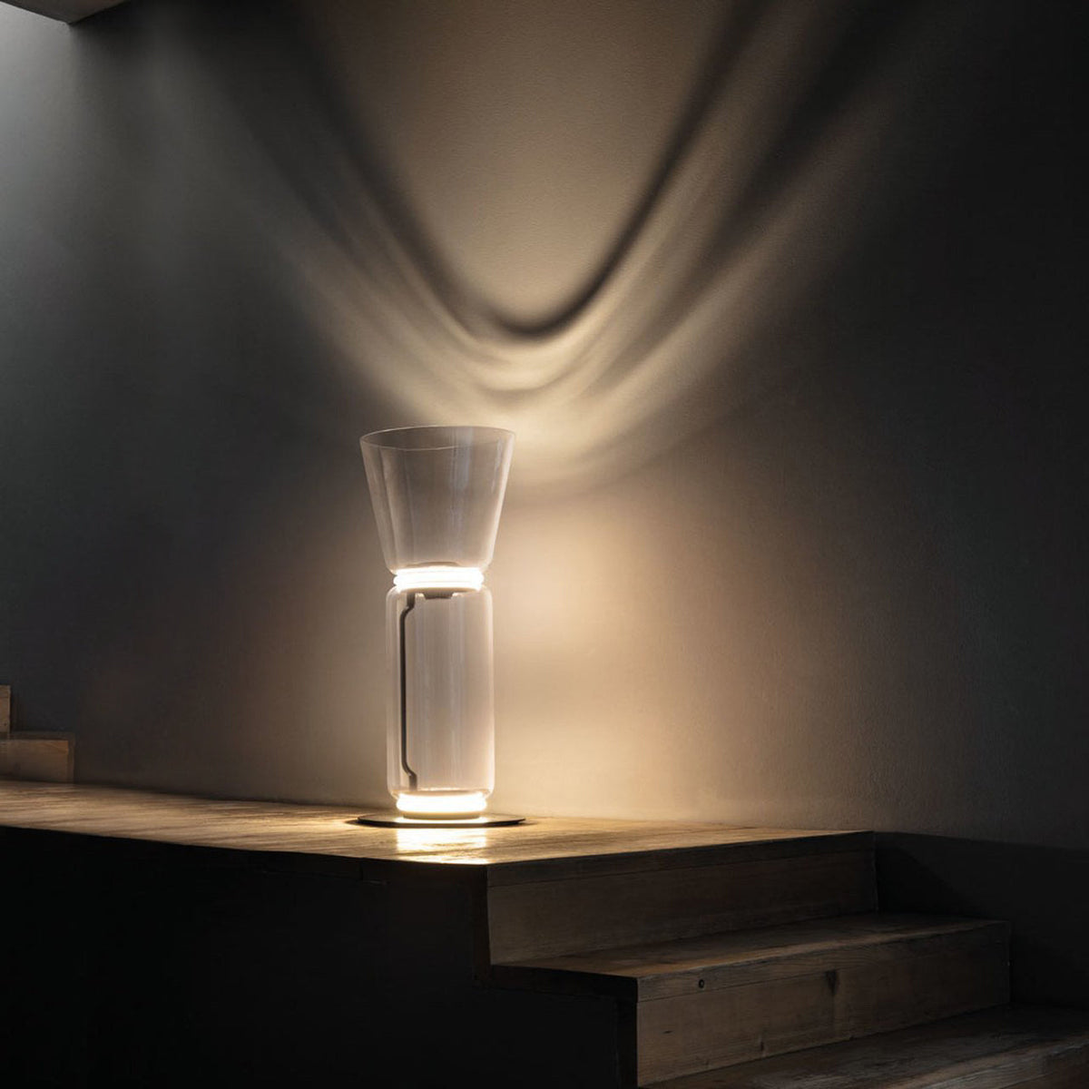 A Clérachic glass floor lamp with a cone shade on a stone step in a dark room, with a fan of warm light on the wall.