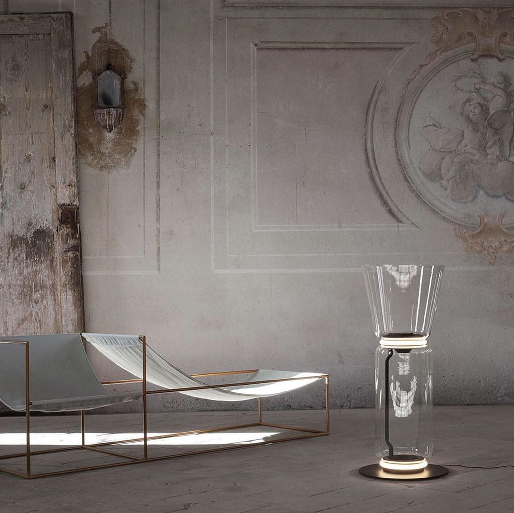A Clérachic glass floor lamp with a cone shade in a room with a plaster relief wall, beside a low lounger with a copper-coloured frame.