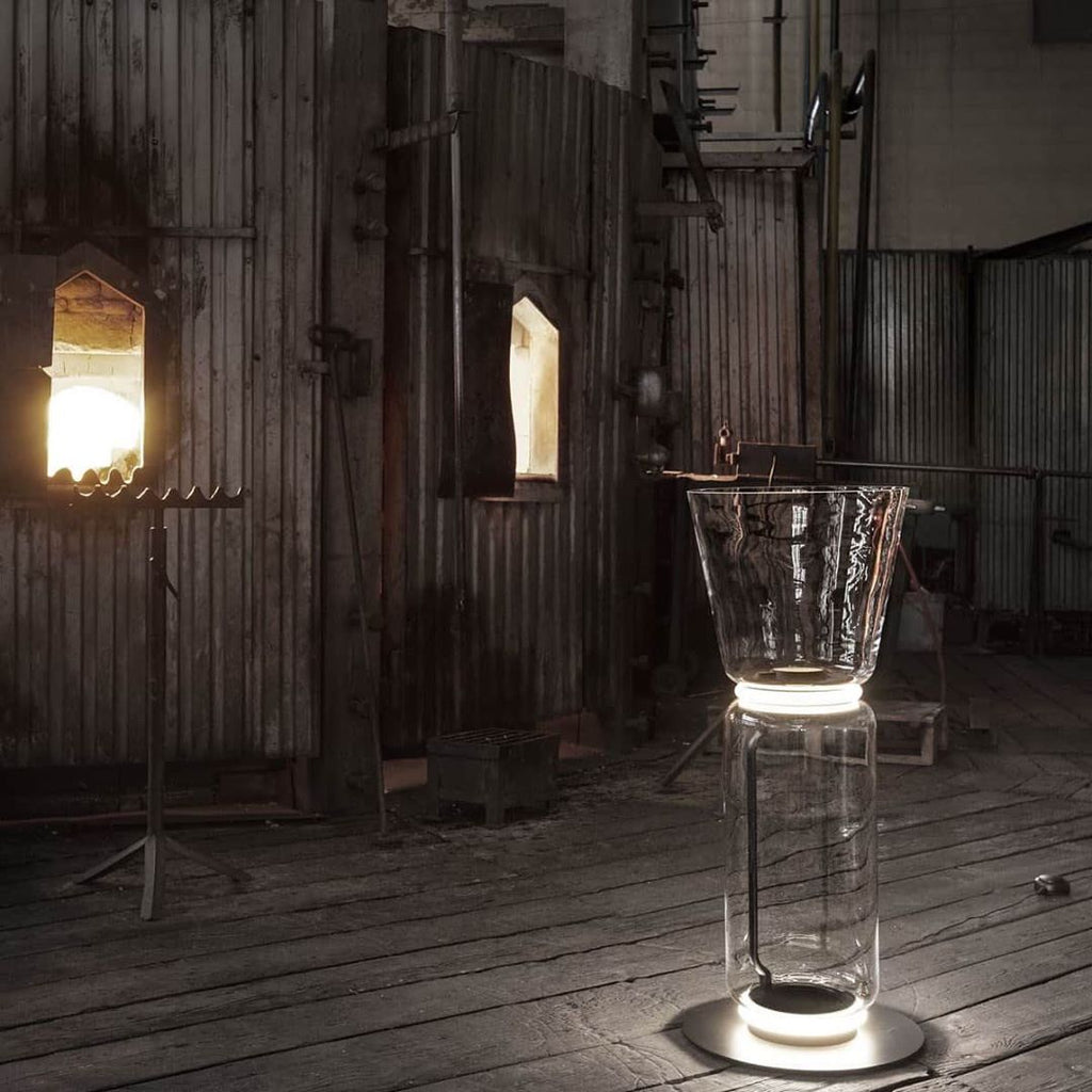 Clérachic glass floor lamp with a cone shade on a plank floor in a timber-walled space, with a lit window opening behind it.