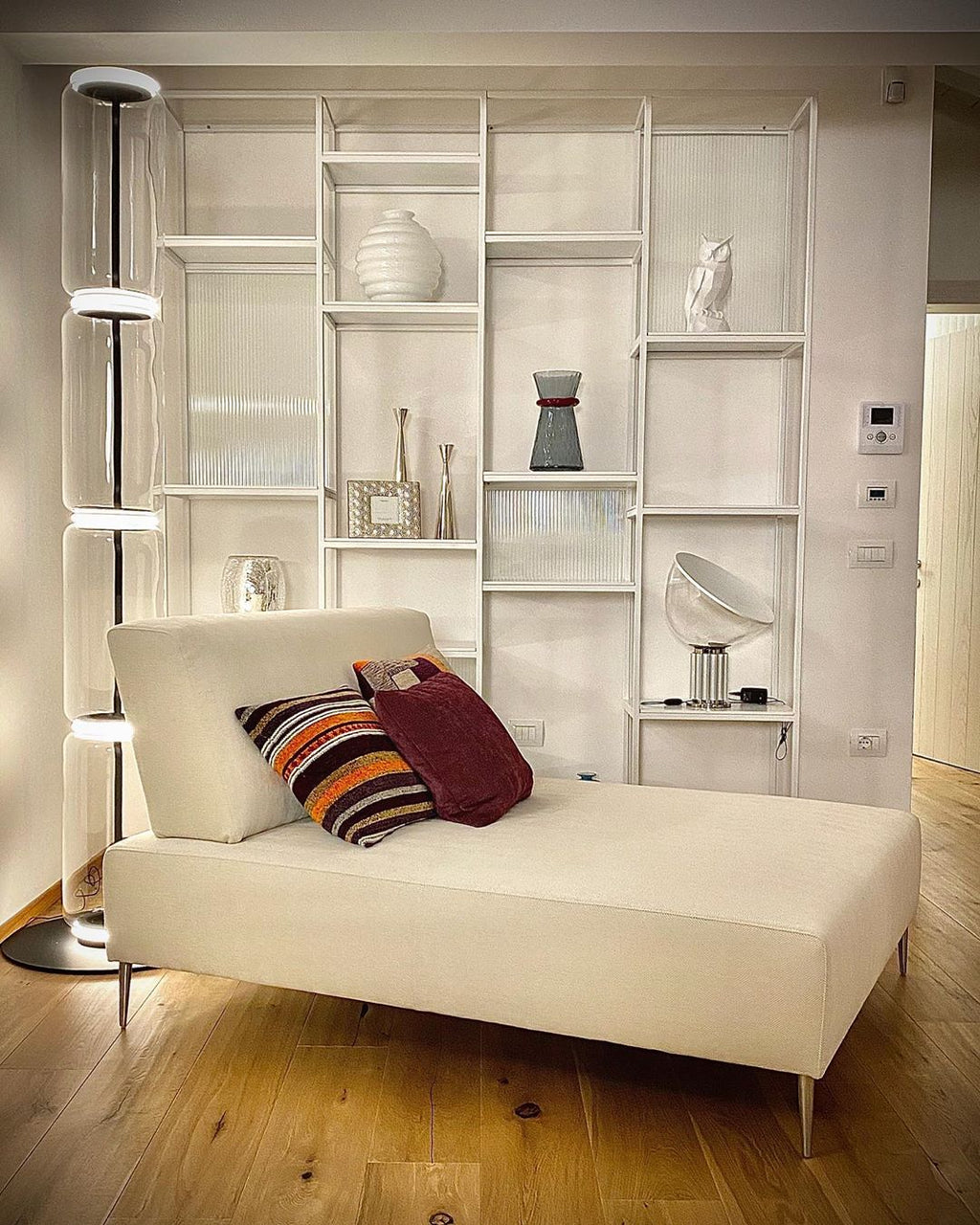 Clérachic glass floor lamp with lit rings, beside a cream chaise longue and a white shelving unit with ornaments.