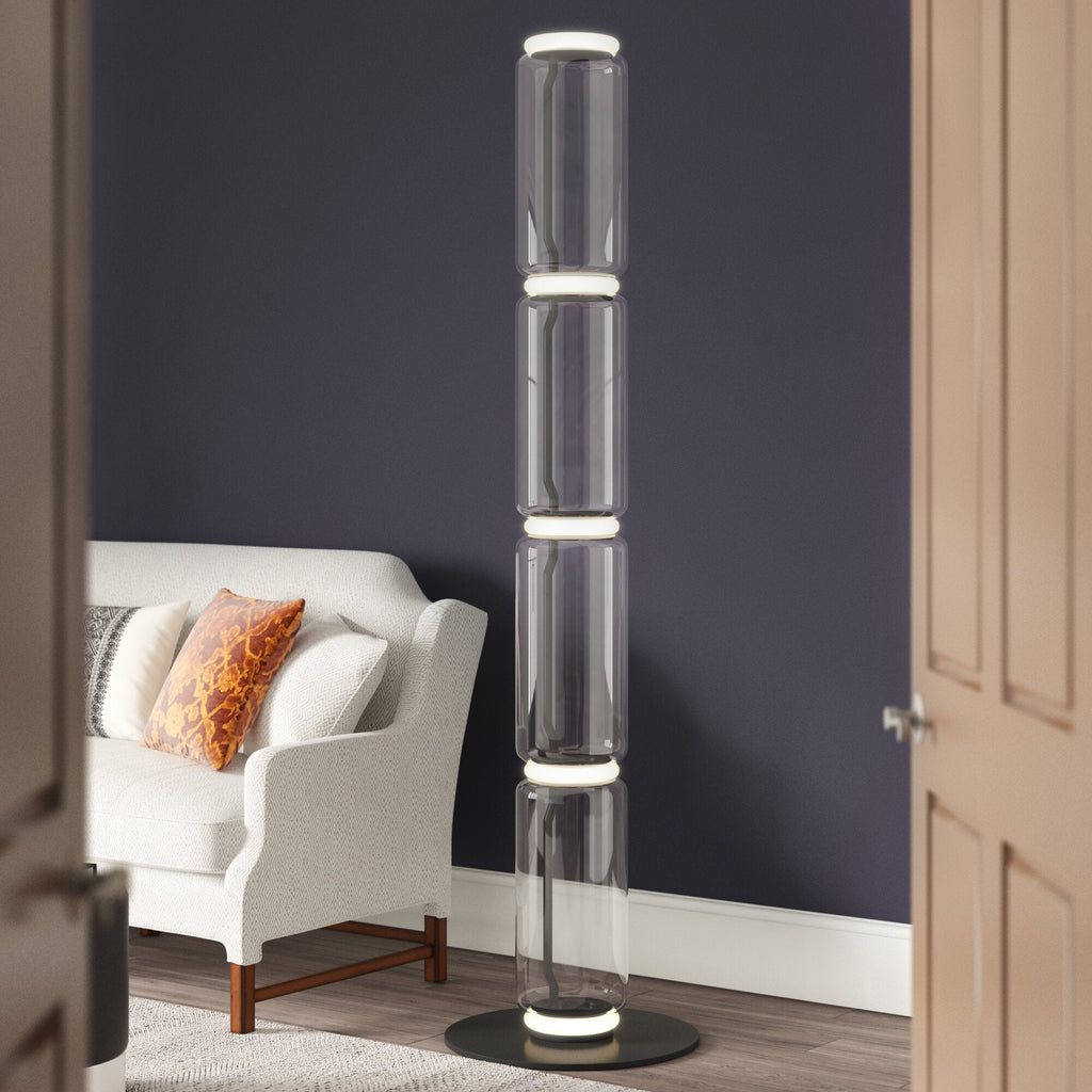 Tall Clérachic glass floor lamp glowing at its rings beside a navy wall and a white armchair with an orange cushion.