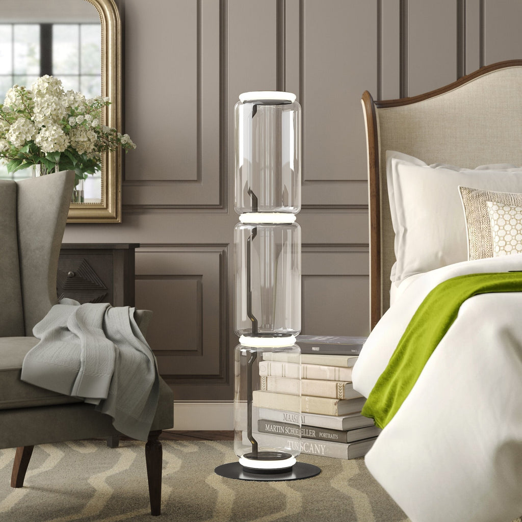 Clérachic glass floor lamp lit in a bedroom with grey panelling, beside a bed, an armchair, white flowers and a stack of books.