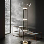 A Clérachic glass floor lamp with a bowl shade, lit and standing beside a copper-framed lounger and a tall narrow window.