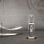 Clérachic glass floor lamp with a cone shade, lit on a stone floor, beside a lounger with a copper frame and a plaster wall.