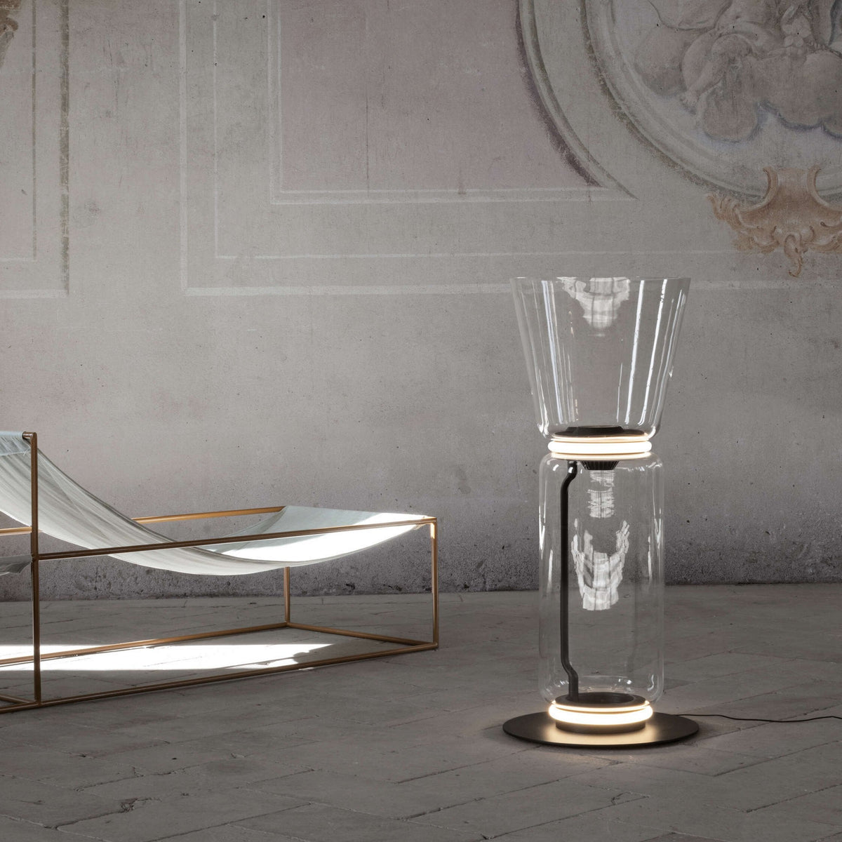 Clérachic glass floor lamp with a cone shade, lit on a stone floor, beside a lounger with a copper frame and a plaster wall.