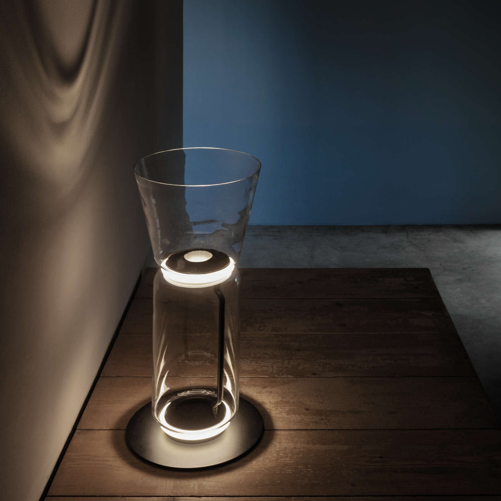 A Clérachic glass floor lamp with a cone shade on a dark wood platform, its base glowing against a blue wall.