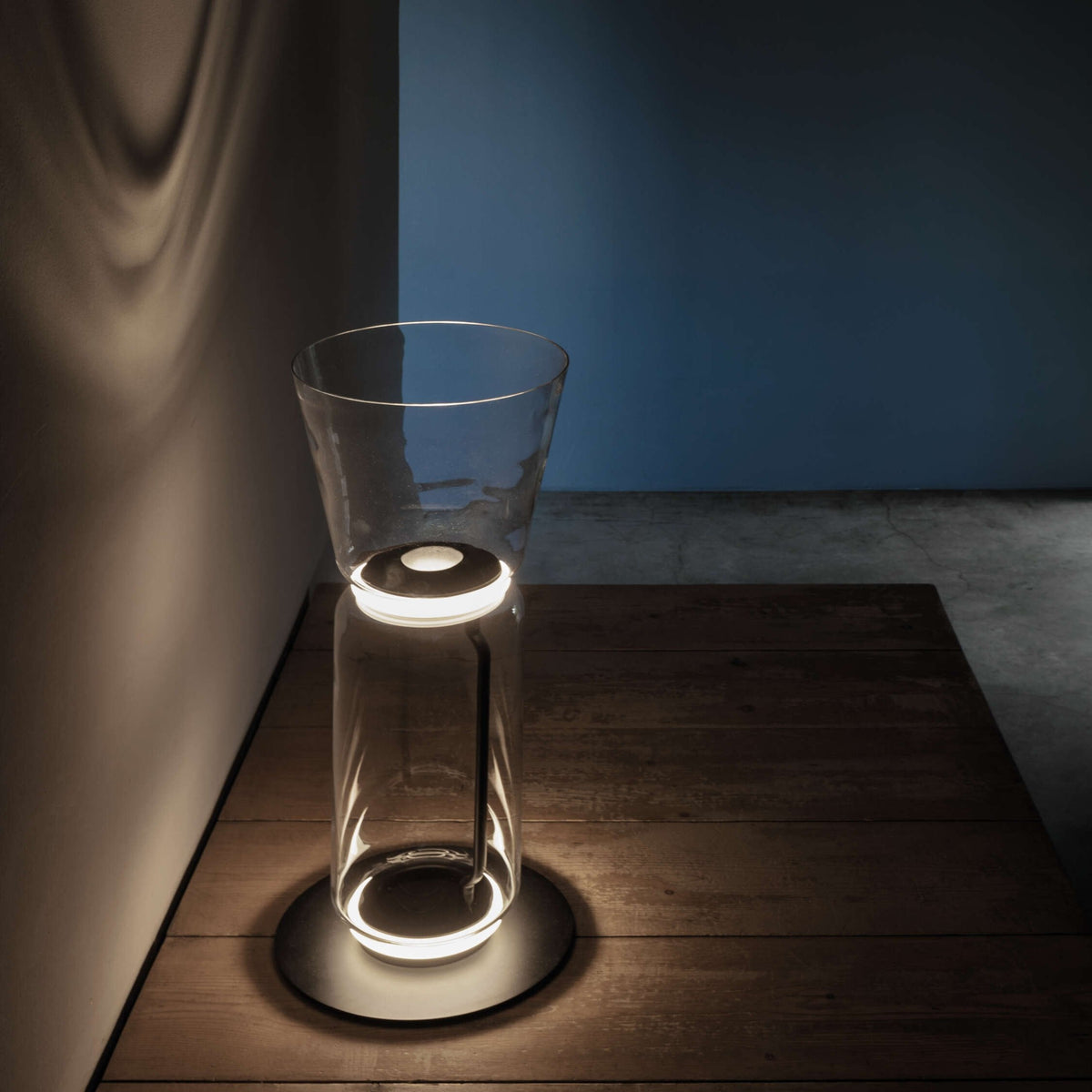 A Clérachic glass floor lamp with a cone shade on a dark wood platform, its base glowing against a blue wall.