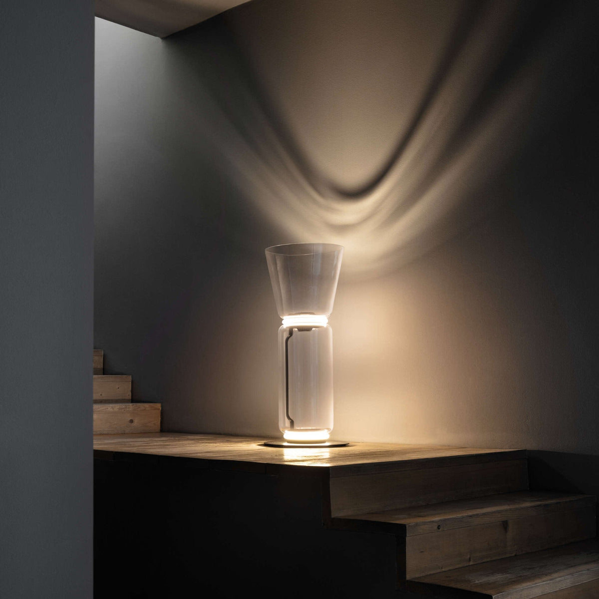 Short Clérachic glass floor lamp with a cone shade on a wooden step, throwing a wave of warm light up a dark wall.