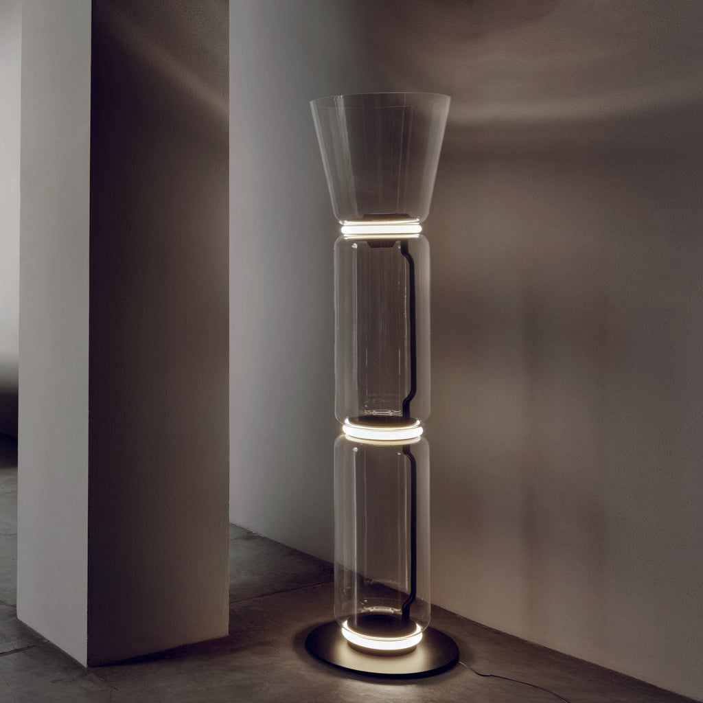 Clérachic glass floor lamp lit at the joints beside a plain pillar, with a cone shade, a stone floor and a warm-grey wall.