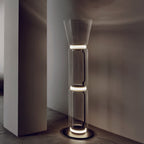 Clérachic glass floor lamp lit at the joints beside a plain pillar, with a cone shade, a stone floor and a warm-grey wall.