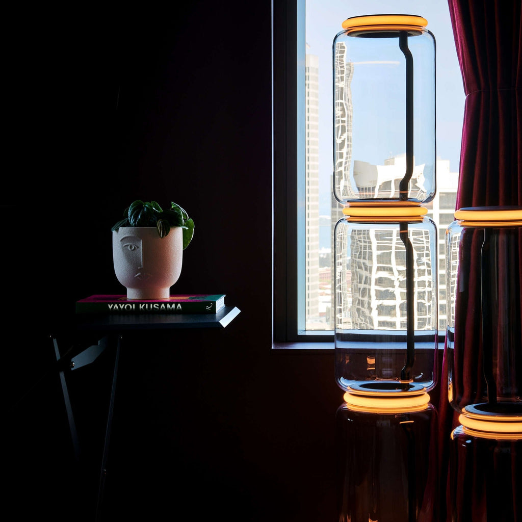 Glass modules of the Clérachic floor lamp with orange-lit rings in front of a city view, beside a red curtain and a plant on a book stack.
