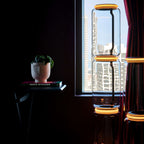 Glass modules of the Clérachic floor lamp with orange-lit rings in front of a city view, beside a red curtain and a plant on a book stack.