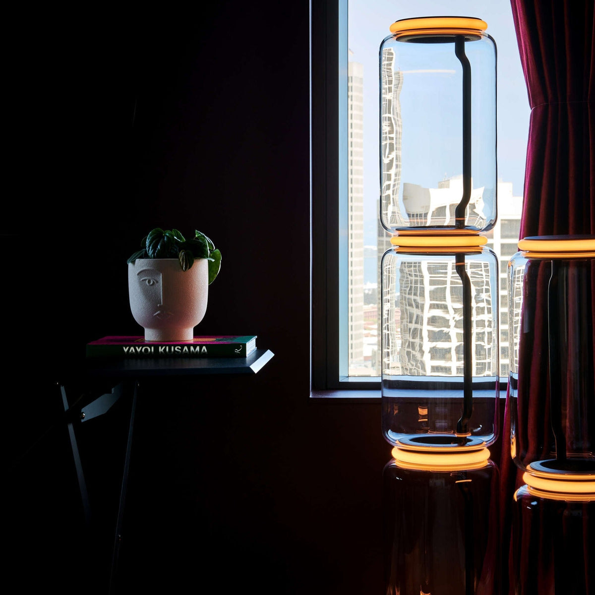 Glass modules of the Clérachic floor lamp with orange-lit rings in front of a city view, beside a red curtain and a plant on a book stack.