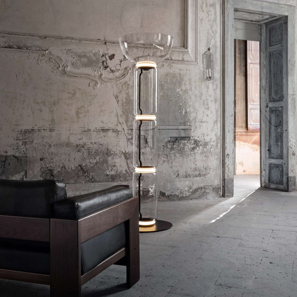 A Clérachic glass floor lamp with a bowl shade in a hall with ornate walls, beside a dark leather and wood sofa and an open doorway.