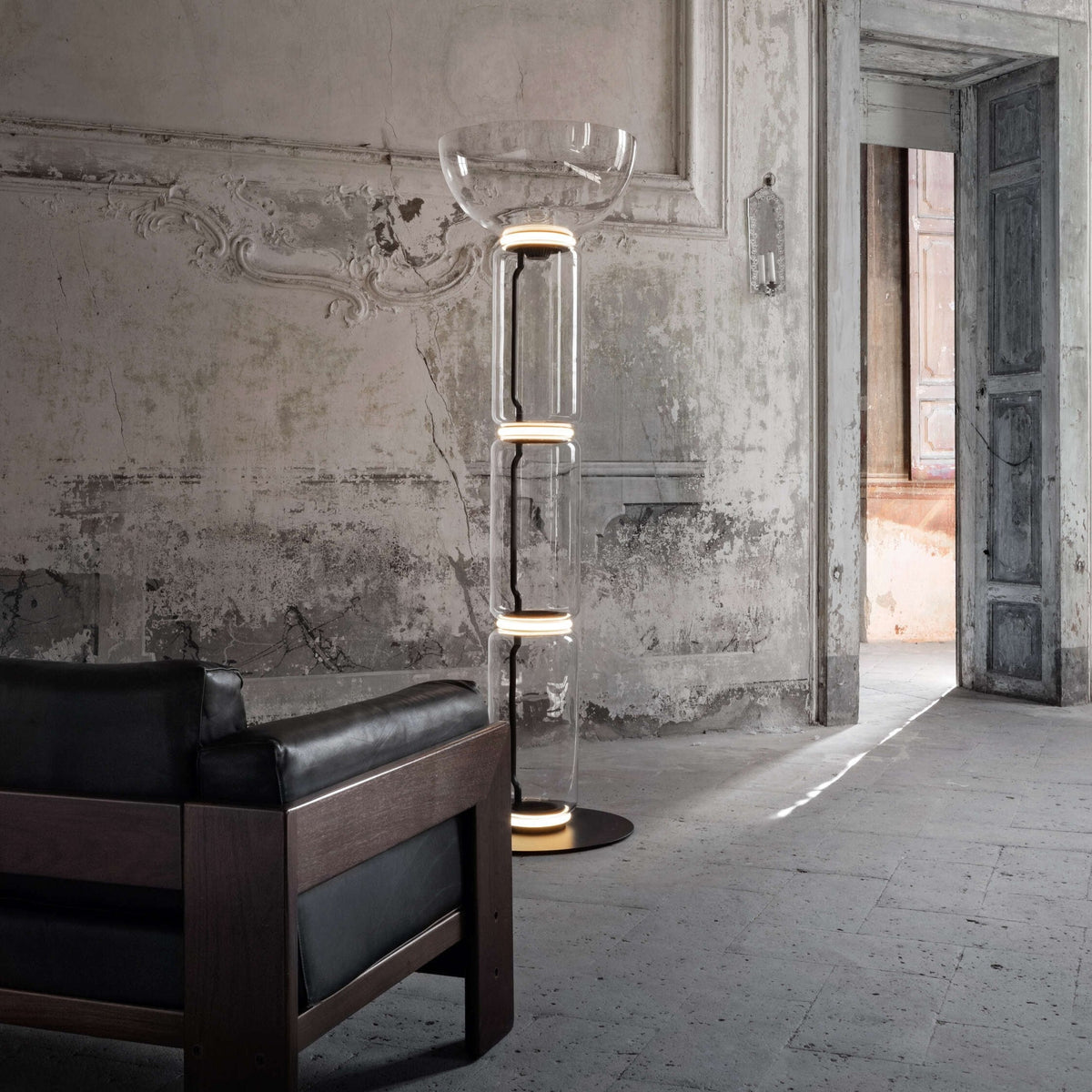 A Clérachic glass floor lamp with a bowl shade in a hall with ornate walls, beside a dark leather and wood sofa and an open doorway.