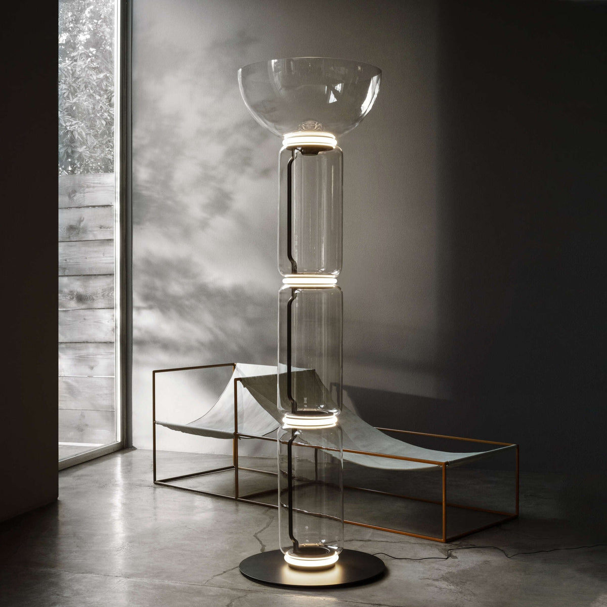 Full-height Clérachic glass floor lamp with a bowl shade, lit beside a lounger with a copper frame and a window onto leaves.