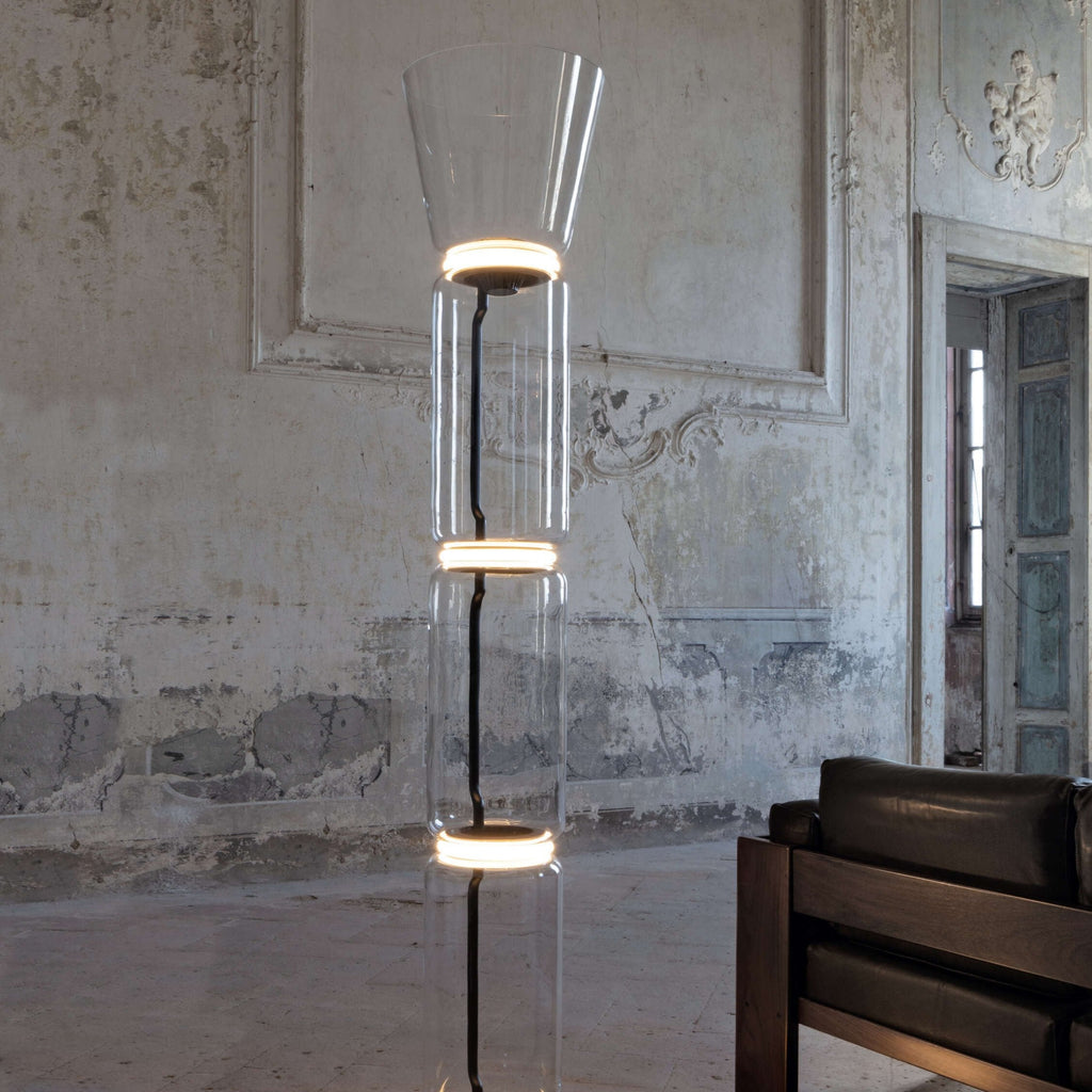 Close crop of this Clérachic glass floor lamp, lit rings at the joints of clear glass, with a black stem and a leather armchair.