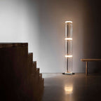 Clérachic glass floor lamp lit in a room with a rough timber staircase and a wooden bench, its glow on the stone floor.