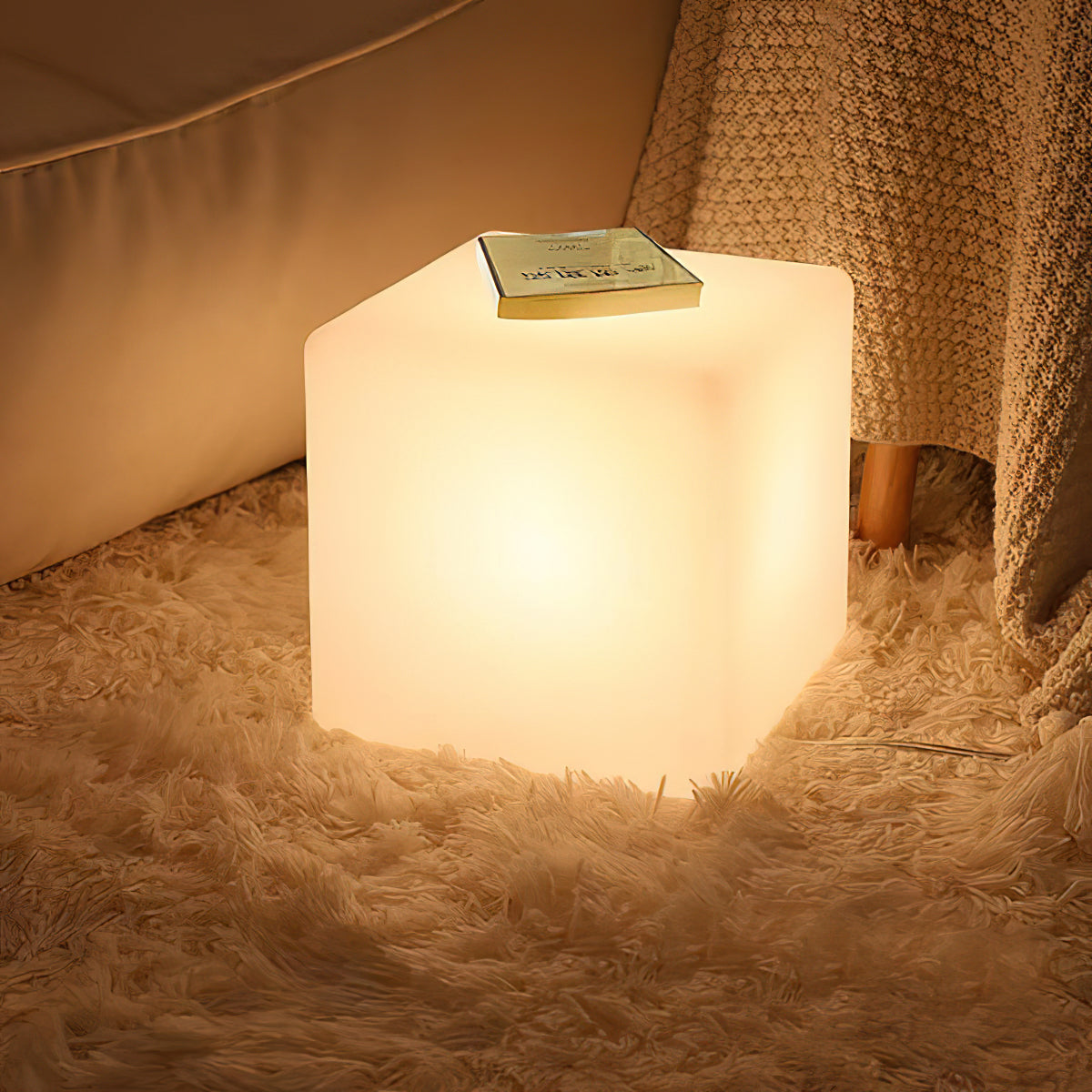 A lit white Clérachic cube lamp on a shaggy rug, with a book on top and a knitted throw draped nearby.