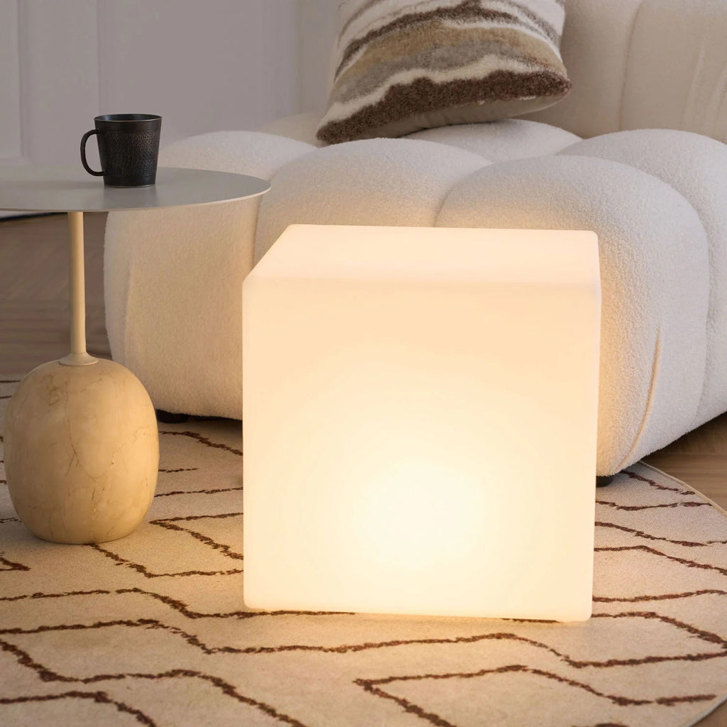 A large lit white Clérachic cube lamp on a zigzag-pattern rug, beside a round metal side table and a bouclé sofa.