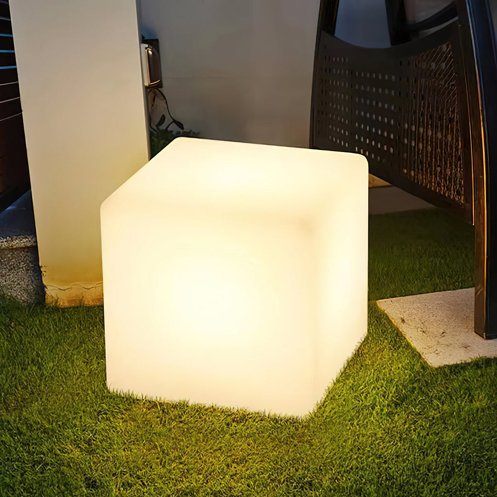 On a patch of lawn beside a dark wicker chair and a stone slab, this white Clérachic cube lamp glows warm.