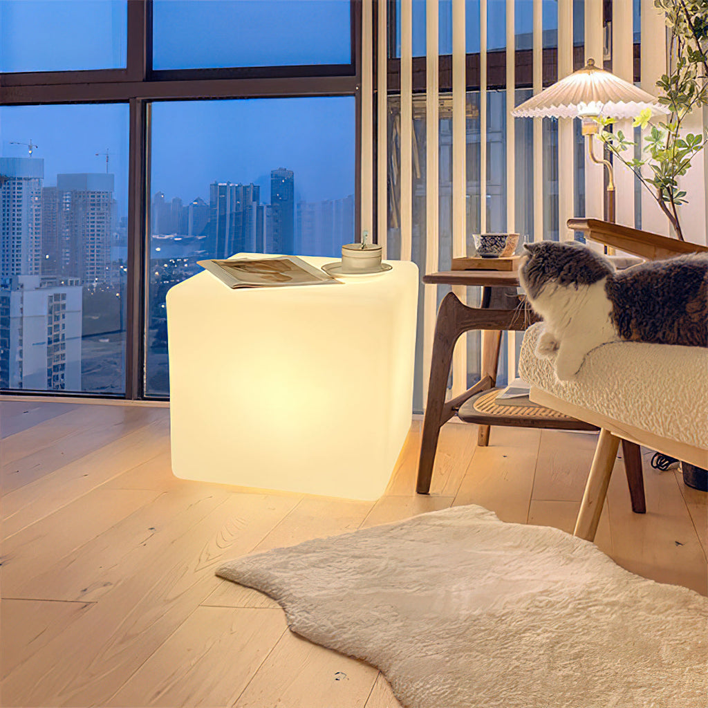 A lit white Clérachic cube lamp in a high-rise living room at dusk, with a magazine on top and a cat on a bench nearby.