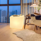 A lit white Clérachic cube lamp in a high-rise living room at dusk, with a magazine on top and a cat on a bench nearby.
