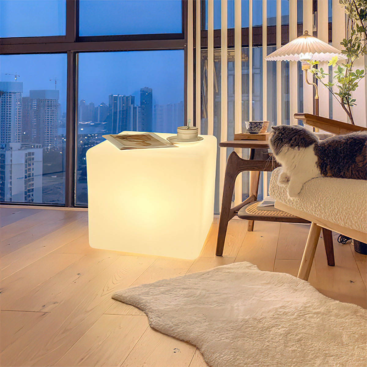 A lit white Clérachic cube lamp in a high-rise living room at dusk, with a magazine on top and a cat on a bench nearby.