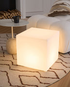 A glowing white Clérachic cube lamp seen from the side, on a patterned rug beside a bouclé sofa and a side table.
