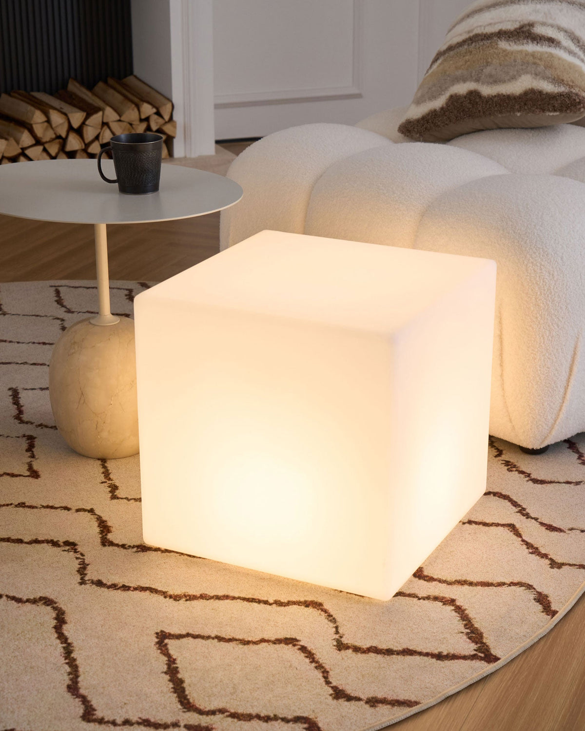 A glowing white Clérachic cube lamp seen from the side, on a patterned rug beside a bouclé sofa and a side table.