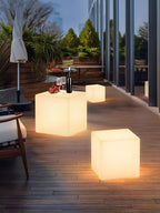 Two lit white Clérachic cube lamps of different sizes on a timber balcony, with a bottle and glasses on the larger one.