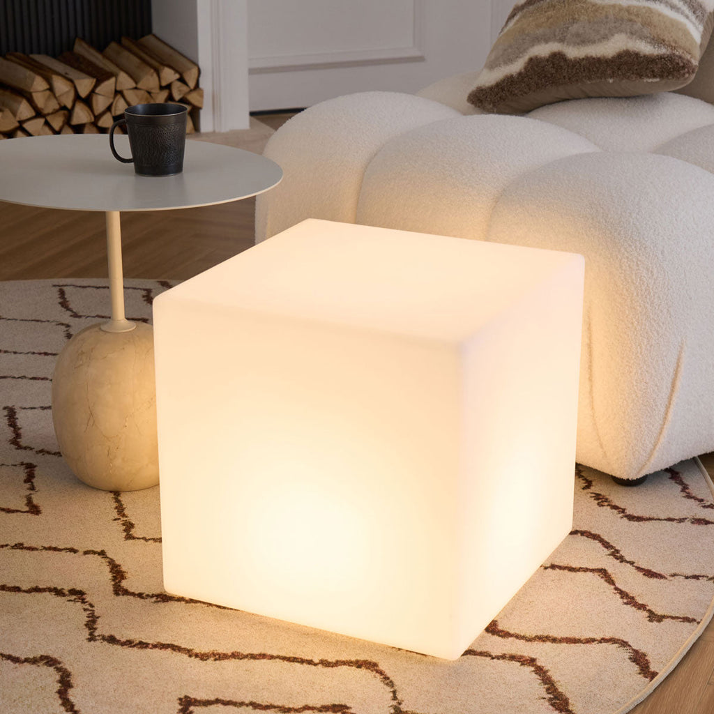 A large white Clérachic cube lamp, lit warm, close beside a cream bouclé sofa and a round side table with a black mug.