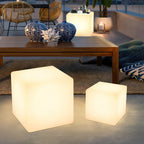 Two lit white Clérachic cube lamps of different sizes on a terrace rug in front of a wooden coffee table with blue vases.