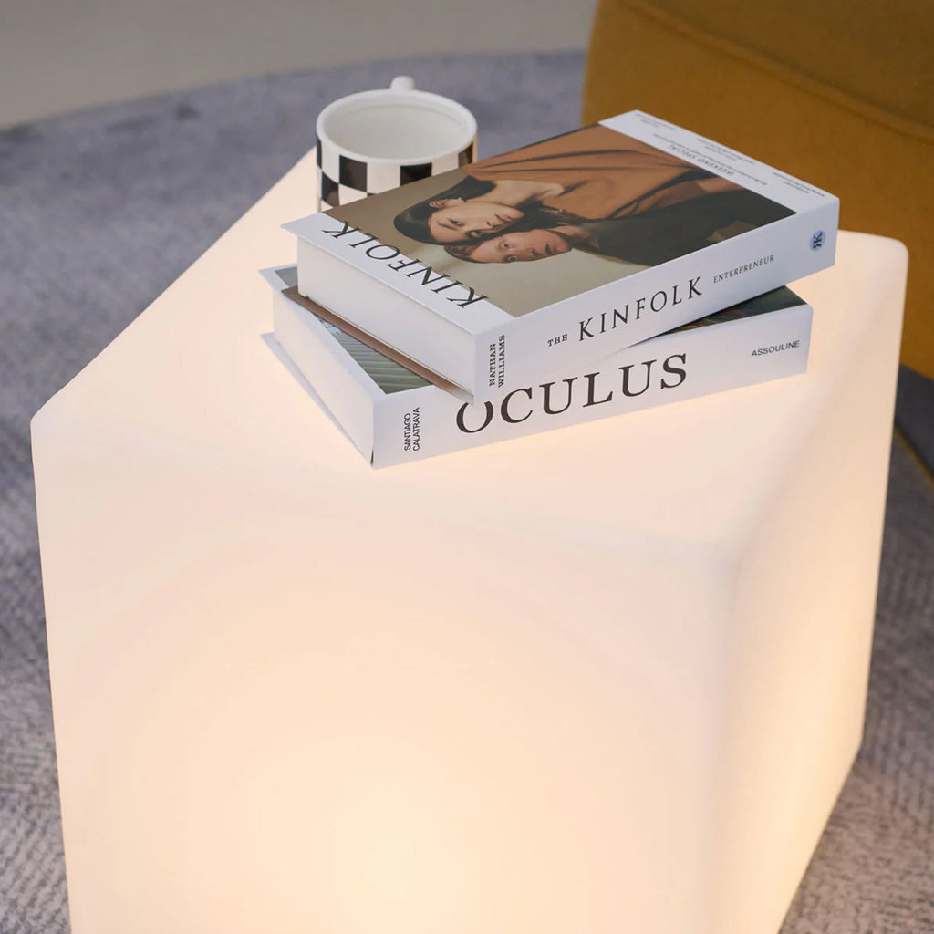 Close view of the top of a lit white Clérachic cube lamp, with a stack of books and a chequered mug resting on it.