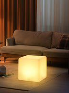 A small lit white Clérachic cube lamp in warm light on the floor, in front of a brown sofa and amber curtains.