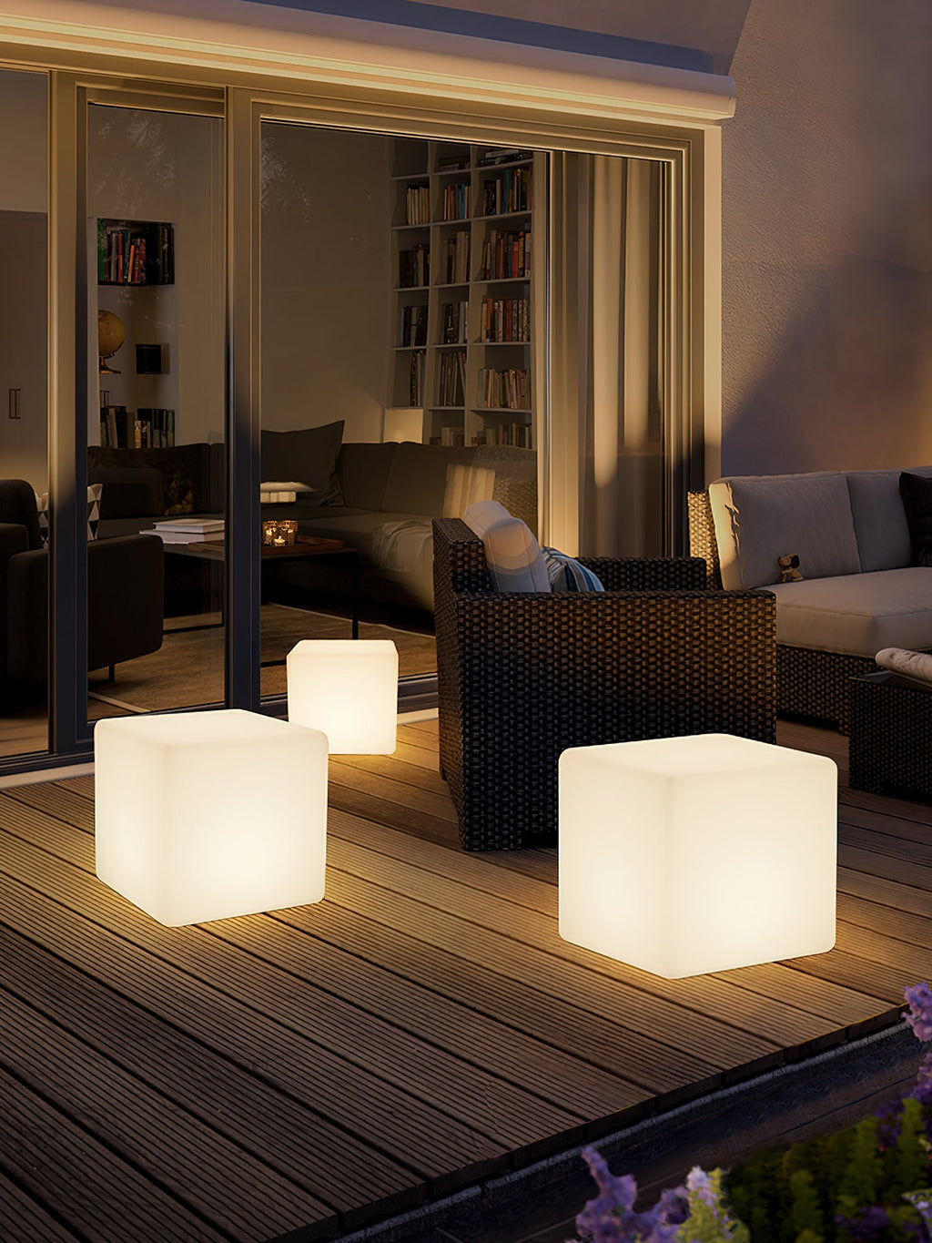 Three lit white Clérachic cube lamps in different sizes on a timber deck at dusk, in front of a glazed living room.